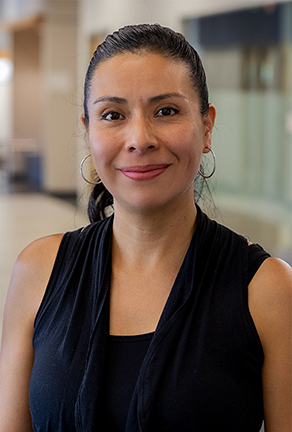 MCC alumna Paola Lintner, with her dark hair pulled back, hoop earrings, and a sleeveless black top, offers a gentle smile in a contemporary indoor environment.