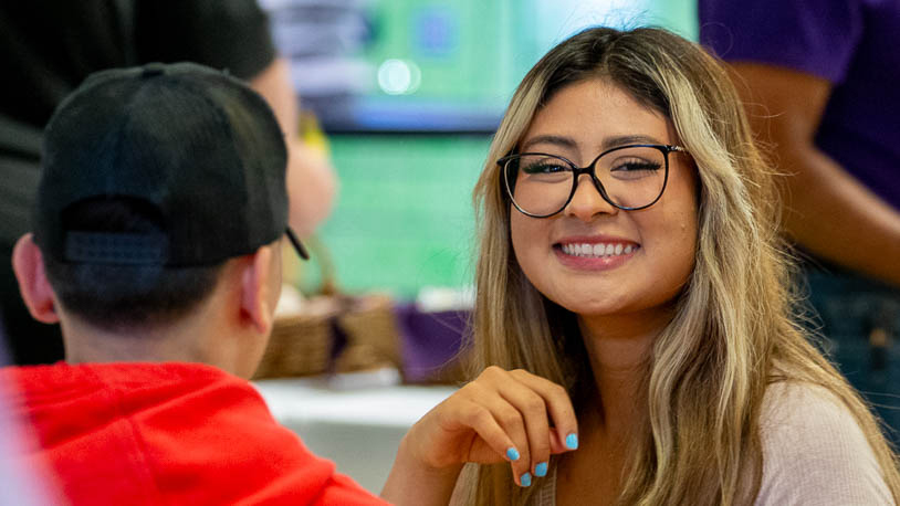 Two MCC students are talking in a casual indoor setting, one wearing a red hoodie and black cap, the other with long blonde hair and blue nail polish.
