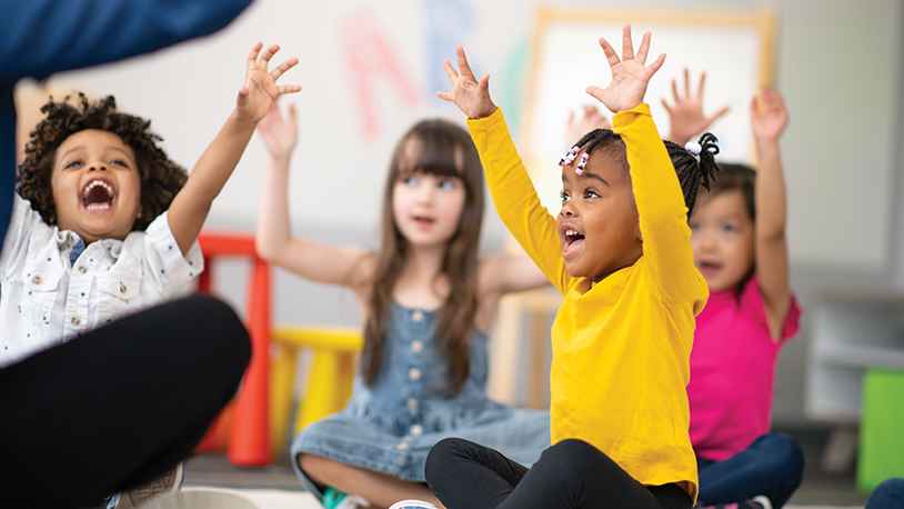 four diverse smiling toddlers seated on the floor raise their hands in the air