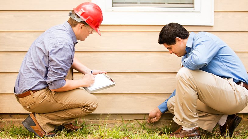 Two White males kneel as they look at the foundation of a house. One man is taking notes on a clipboard.