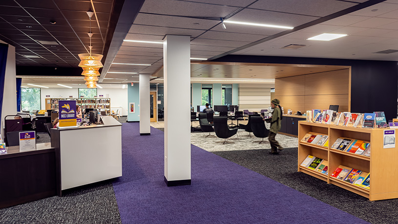 Modern library interior with purple carpet, wooden bookshelves displaying colorful books, and a person walking near computer workstations.