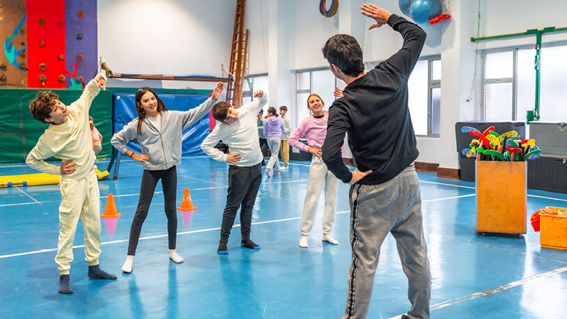 A teacher faces a gym class of four smiling middle school kids as he leads them in stretching.