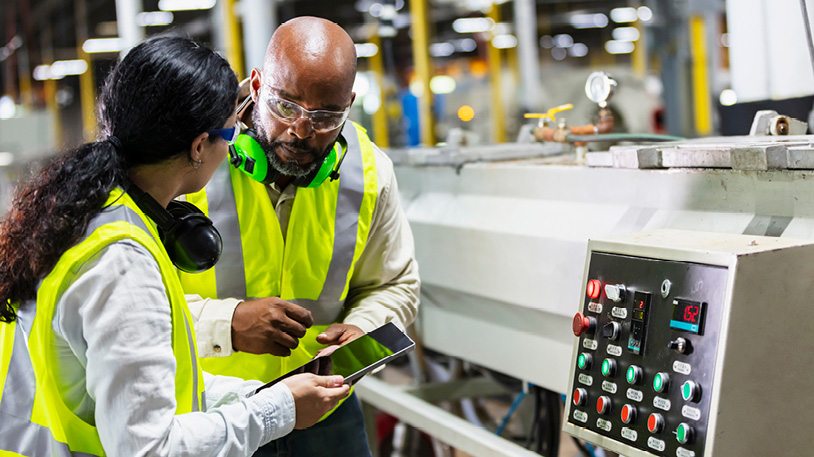 Two manufacturing workers in safety vests and goggles discussing data on a tablet near industrial machinery controls.