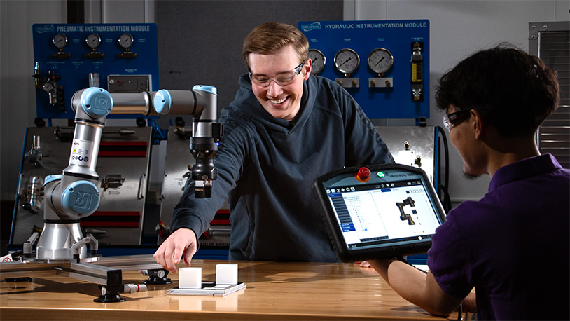A White male MCC student in his early 20's smiles as he reaches across a table to move something under a robotic arm. Another student has a tablet computer.