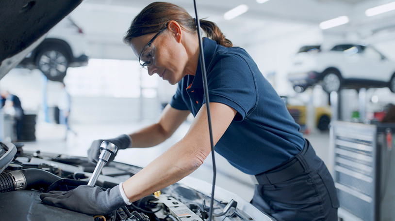 Female mechanic leans over engine bay of car and uses a ratchet while wearing gloves