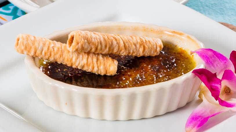 A crème brûlée dessert topped with two wafer cookies, served in a ramekin on a white plate with an orchid garnish.