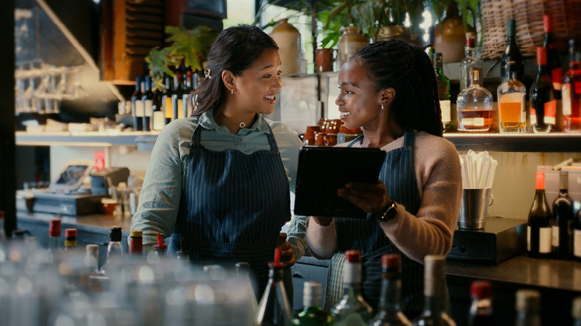 Two smiling bartenders wearing aprons collaborating while looking at a tablet behind a counter stocked with various bottles