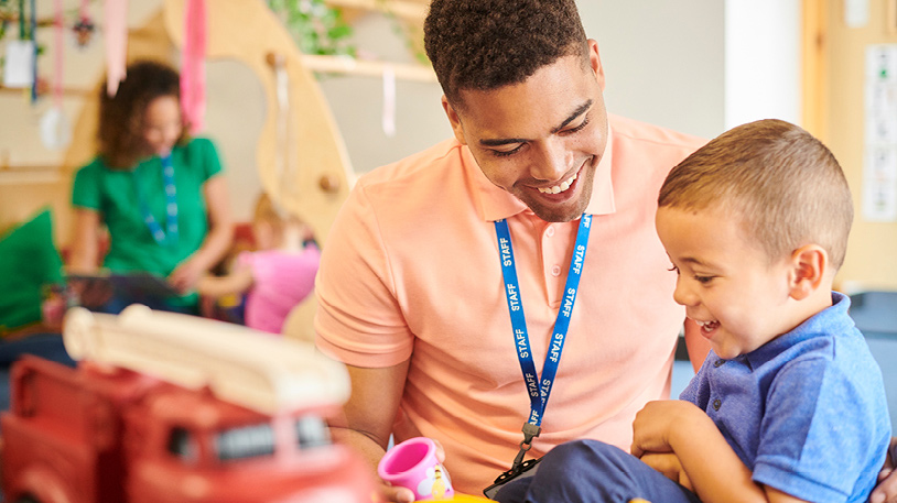 Smiling young male teacher engaging happily with a laughing boy during playtime in a colorful classroom setting.