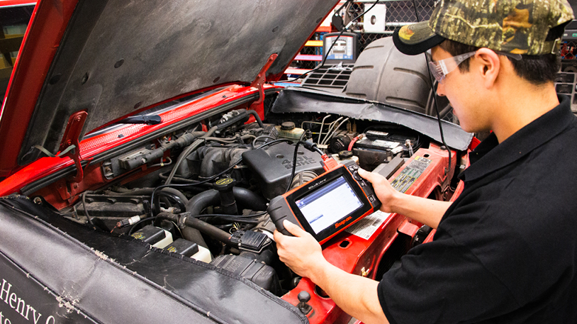Mechanic student stands in front of a truck with its hood open. Student uses a scan tool tablet to diagnose an issue with the truck engine