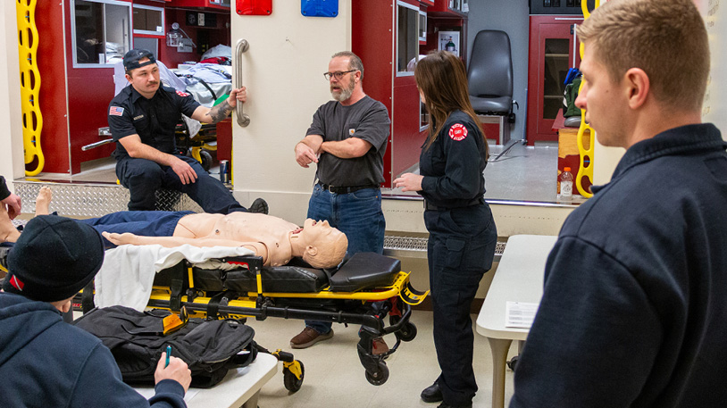 MCC students gathered around a medical training dummy on a stretcher inside the simulated ambulance bay during a CPR demonstration.