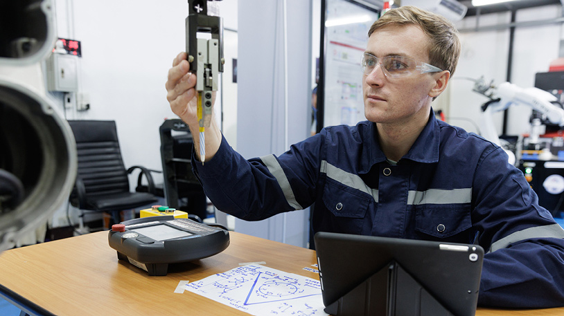 An engineer in safety glasses inspecting a mechanical component in a lab with technical drawings and a tablet on the desk.