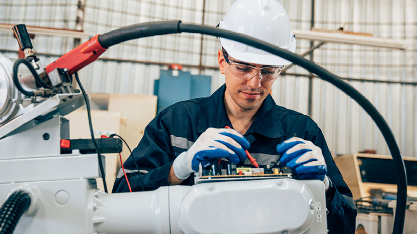 A technician wearing safety gear is repairing a robotic arm in an industrial workshop.