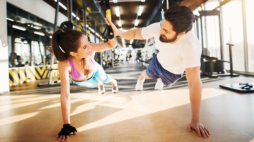 A man and woman in athletic wear doing side-by-side plank exercises while giving each other a high five in a bright gym.
