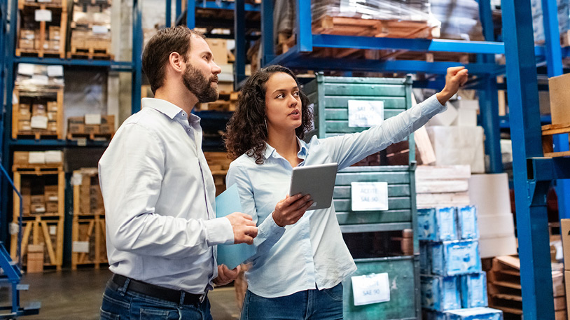 Two warehouse workers discuss inventory details while reviewing a tablet in a storage area filled with stacked boxes and pallets.