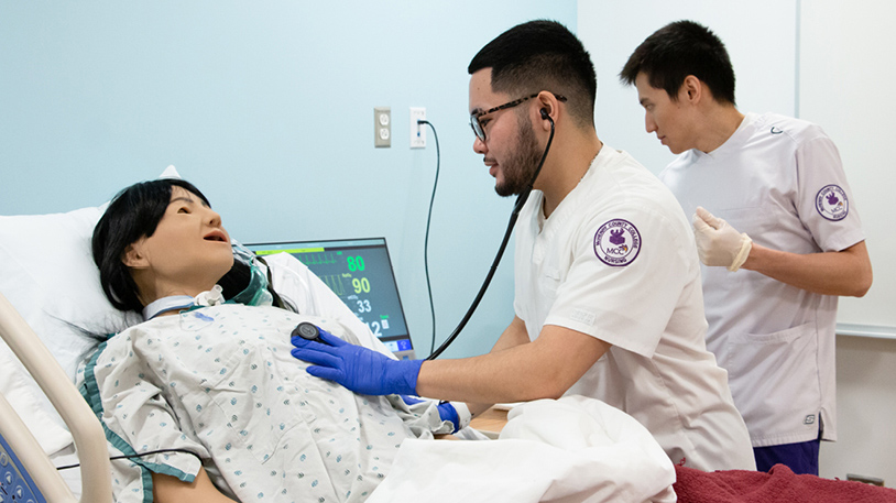 An MCC student uses a stethoscope to examine a patient mannequin in a clinical training setting.