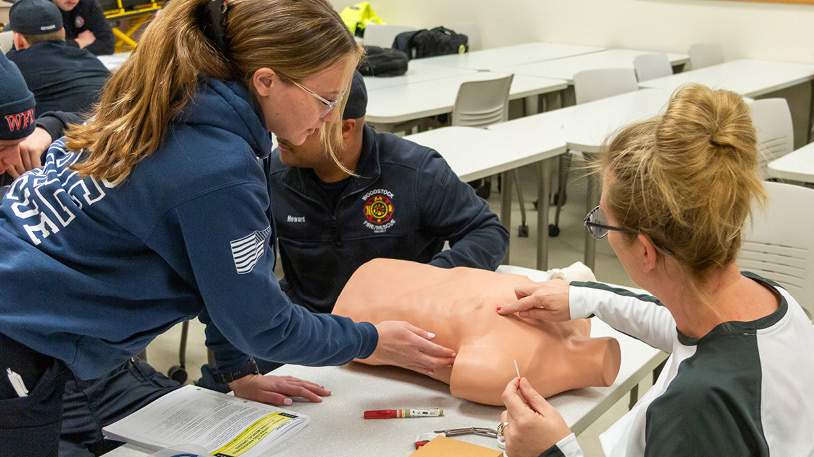 Two women practicing medical procedures on a CPR training mannequin in a classroom setting.