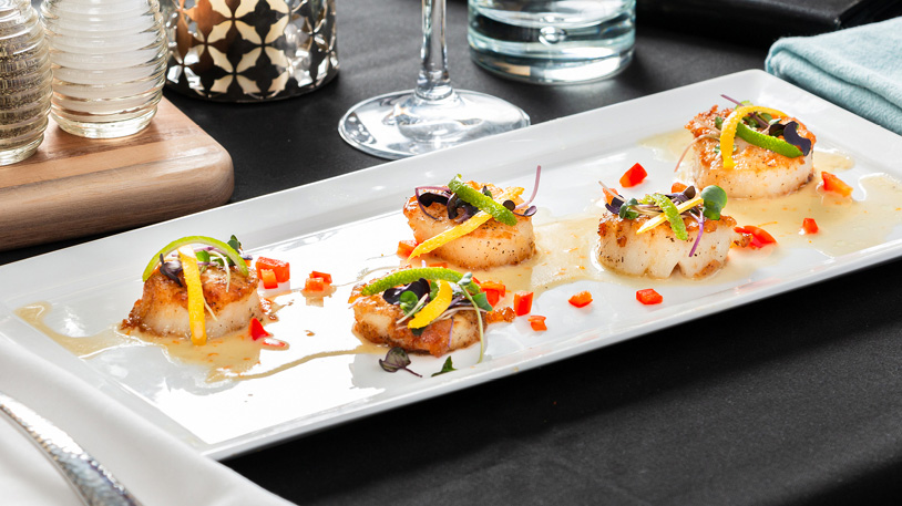 Five seared scallops garnished with microgreens, thinly sliced lemon, and diced red peppers on a rectangular white plate with a light sauce.