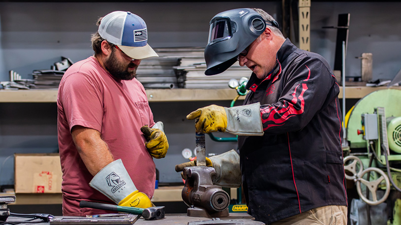 Two men in a workshop, one wearing a welding helmet and gloves, working on a metal piece clamped in a vise, the other observing closely.