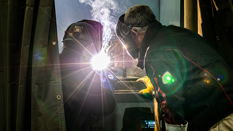 Two workers wearing protective gear are welding, producing bright sparks and smoke in an industrial setting.
