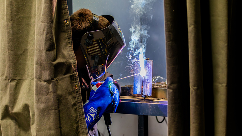 Welder wearing protective gear and helmet, welding metal pieces with sparks and smoke visible.