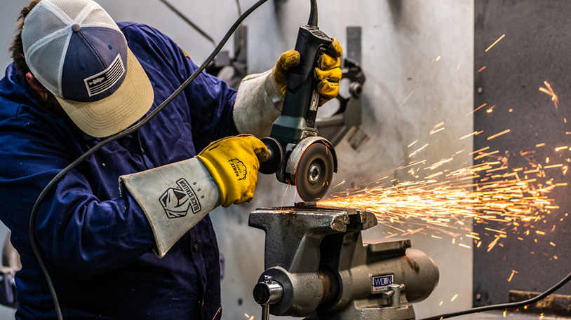 A worker wearing protective gloves and a cap is using an angle grinder to cut metal, creating bright sparks in a workshop.