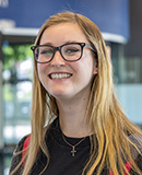 MCC alumna Sammie Crager, with long blonde hair and glasses, smiles indoors by a blue wall and glass panels.