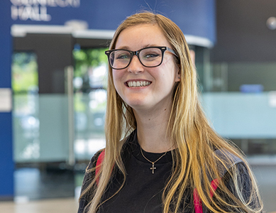 MCC alumna Sammie Crager, with long blonde hair and glasses, smiles indoors by a blue wall and glass panels.