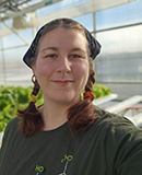 MCC horticulture student Jyinxs Crisp is wearing a dark green shirt and bandana, standing inside MCC's greenhouse with rows of hydroponically grown leafy green plants.