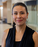 MCC alumna Paola Lintner, with her dark hair pulled back, hoop earrings, and a sleeveless black top, offers a gentle smile in a contemporary indoor environment.