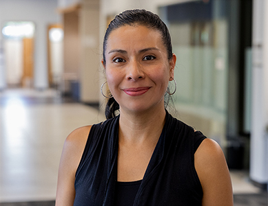 MCC alumna Paola Lintner, with her dark hair pulled back, hoop earrings, and a sleeveless black top, offers a gentle smile in a contemporary indoor environment.