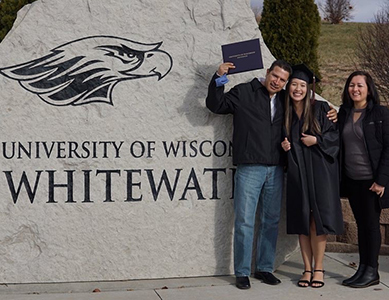 MCC alumna Carina Lopez  is wearing her graduation cap and gown, standing with her parents in front of a rock that says University of Wisconsin White Water