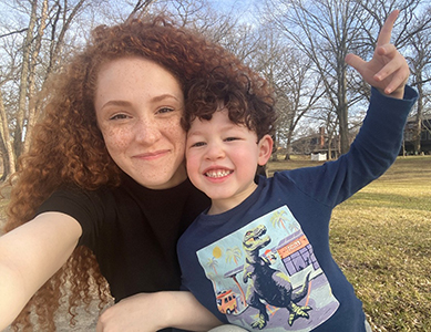 MCC alumna Katherine Sanders smiles as she hugs a cheerful young boy wearing a dinosaur-print sweatshirt outdoors on a sunny day.