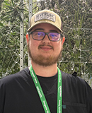 MCC alumnus Nate Voorhees, wearing a beige Neversink Farms cap and a black shirt, stands in front of tall cannabis plants inside a greenhouse.