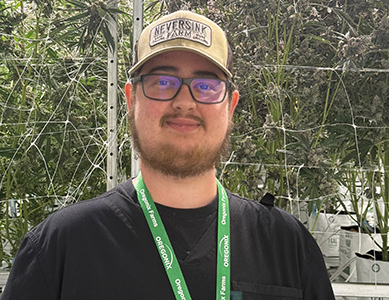 MCC alumnus Nate Voorhees, wearing a beige Neversink Farms cap and a black shirt, stands in front of tall cannabis plants inside a greenhouse.