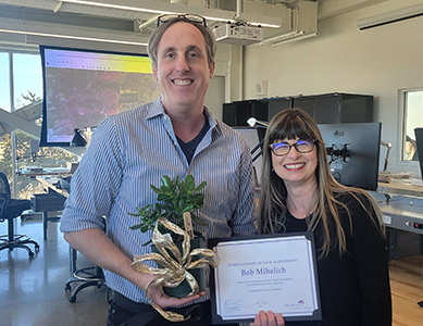 Bob Mihelich is holding a small potted plant. He is standing next to Daniela Broderick, who is holding a certificate of recognition