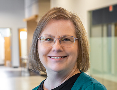  Jean Gaspari, coordinator of the University Center at MCC, a middle-aged woman with short blonde hair and glasses, wearing a teal cardigan over a black top in a modern indoor setting.